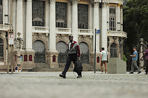 Man walking across a cobblestone plaza in front of a grand historic building with arched windows and columns, carrying a backpack; other people and street signs in the background.