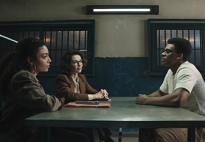 Duas mulheres, as atrizes Marjorie Estiano e Any Gabrielly, se sentam com um homem, o ator Hypólito, dentro de uma penitenciária
