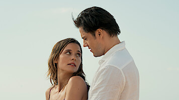 A woman and man standing close together on a beach, looking into each other's eyes with a clear sky in the background.