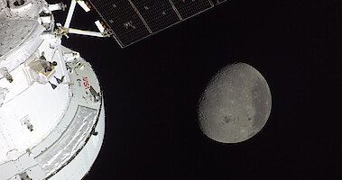 Part of a spacecraft in space with solar panels visible, and the moon in the background against the blackness of space.