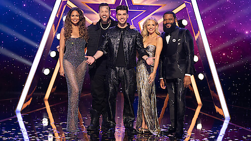 Chrissy Teigen, Jelly Roll, TJ Salta, Sarah Michelle Gellar, and Anthony Anderson dressed in sparkling outfits stand smiling together on a stage with a star-shaped backdrop and dramatic purple lighting, confetti on the floor, and a festive atmosphere