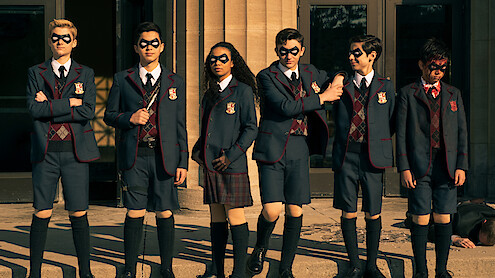 Six children in matching school uniforms and black eye masks stand in a row outside a building with large columns, in sunlight, appearing serious or ready for action. Another person is lying on the ground in the background.