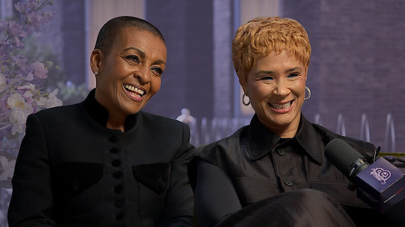 Two women smiling during an interview, seated indoors with a floral arrangement and large windows in the background, both wearing black outfits, with a microphone labeled “20” visible in the foreground.