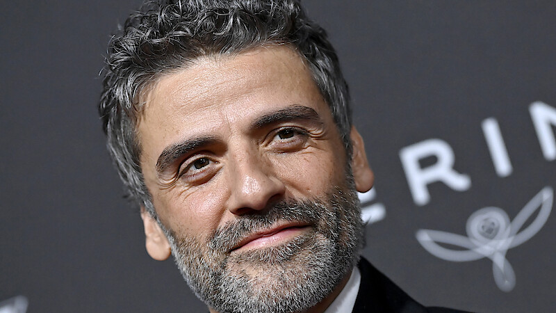 Man with curly hair and beard in formal attire smiles at a black tie event, standing in front of a dark backdrop with white decorative text and floral graphics.
