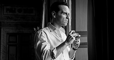 Black and white photo of Andrew Scott as Tom Ripley holding a camera while looking out of a window in Season 1 of 'Ripley'