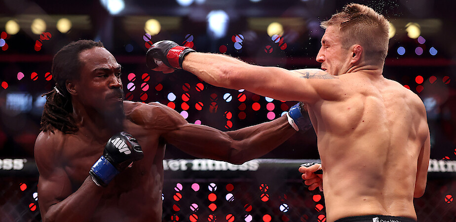 Two male MMA fighters in a ring, one landing a punch to the other’s face during a match, under bright arena lights with a blurred audience and cage fence in the background.