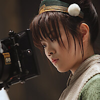 Miyako Cech as Toph on a film set, focused intently on a professional movie camera during a scene, with soft lighting and a blurred studio background.