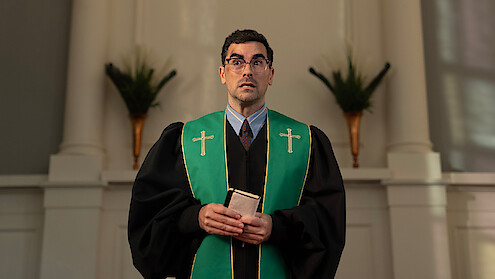 A man dressed in clerical robes with green stole, holding a book, stands in a church sanctuary with white walls, columns, and decorative plants in the background, looking slightly upward.