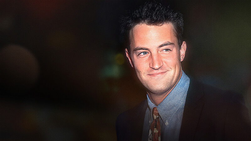Man in a suit and patterned tie smiling, standing against a dark, softly blurred background with gentle ambient lighting, suggesting a formal or evening event.