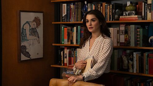 Rachel Weisz as The Protagonist  in a white blouse stands in front of a bookshelf filled with books and decor, holding folders. A framed artwork hangs on the wall, creating a cozy, intellectual office or library setting.