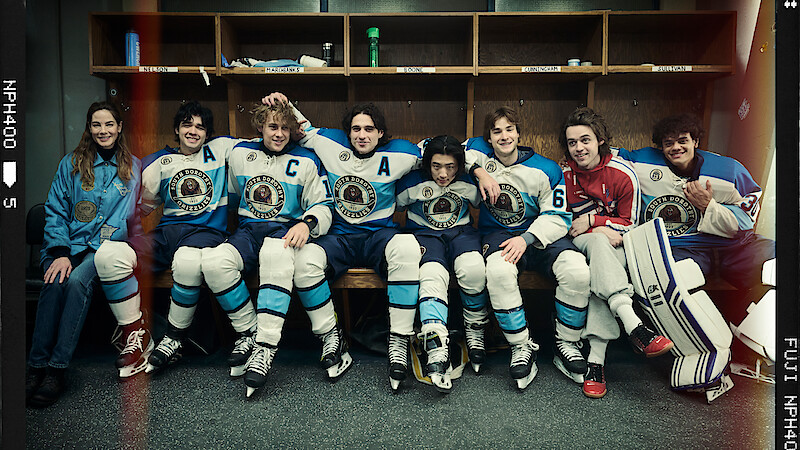 A group of young hockey players in blue and white uniforms and one in red sit together on a locker room bench, posing and smiling for a team photo, hockey gear and sticks by their sides.