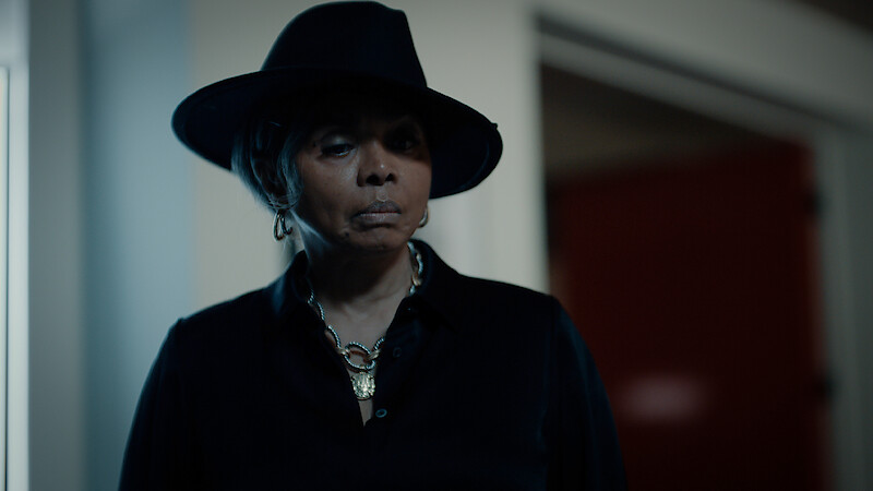 Person wearing a dark wide-brimmed hat and black outfit stands indoors in dim lighting, with a serious expression, silver jewelry visible, and blurred background showing a doorway and red wall.