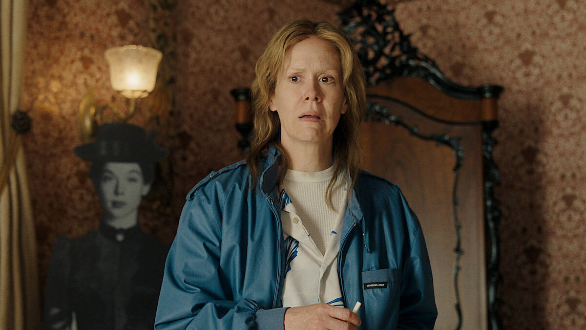 A concerned person in a blue jacket stands in a vintage room with patterned wallpaper, ornate wooden furniture, and a black-and-white cutout of a woman in period clothing in the background.
