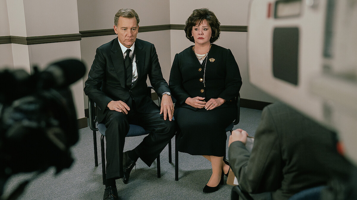 A man and woman in formal attire sit side by side holding hands, appearing serious, in what looks like an interview or televised setting with cameras and a neutral office background.