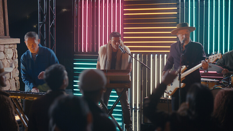 A band performs live on stage with colorful neon lights in the background, including a keyboard player, singer, and guitarist. Audience members are watching and raising their hands in an outdoor nighttime concert setting.