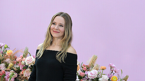 Kristen Bell wearing an off the shoulder black dress in front of a low wall of flowers behind her and a seamless lavender wall at an Los Angeles event for Nobody Wants This Season 2.