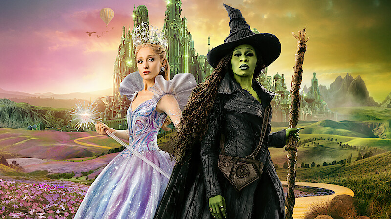 A magical fairy in a sparkling gown and a green-skinned witch with a pointed hat stand in a colorful fantasy landscape with a yellow brick road, flowers, and an emerald city in the background.