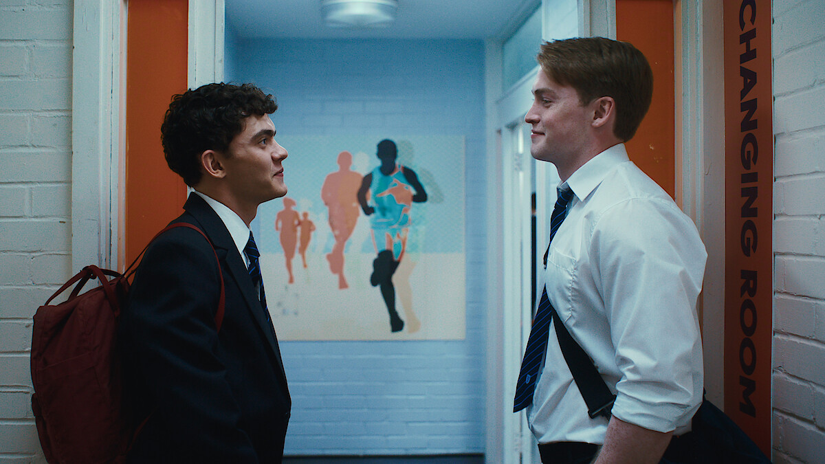 Two students in school uniforms stand facing each other in a hallway near a changing room, with sports bags and a colorful runner-themed painting on the wall behind them. The setting has white brick walls and blue lighting.