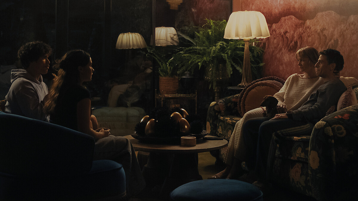 Four people sit across from each other in a dimly lit cozy living room with vintage furniture, lamps, houseplants, and a round coffee table, suggesting an intimate evening conversation.