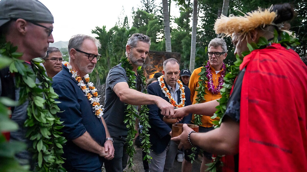 Group of people wearing leis and greenery participate in an outdoor traditional ceremony led by a person in red attire and a feathered headdress, surrounded by tropical trees.