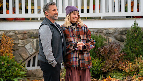 A man and woman stand outdoors in autumn clothing, smiling in front of a stone wall and white railing, surrounded by fall foliage and plants.