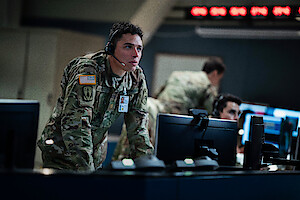 US military officer in camouflage with headset leans over monitor in command center, focused on screens; other soldiers work in background, intense and serious atmosphere.