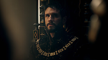 A solemn man in elaborate medieval attire with a gold chain sits on a dark, ornate throne in a dimly lit, regal setting, creating a moody, contemplative atmosphere.