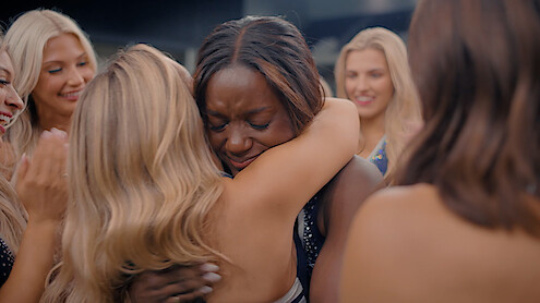 A group of women in a celebratory indoor setting, with one woman hugging another who appears emotional while others smile and clap around them.