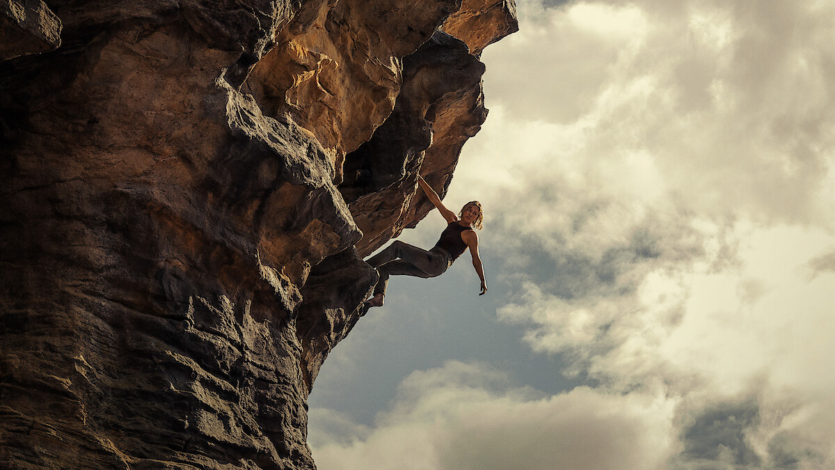 Person rock climbing on a steep, overhanging cliff with dramatic rock formations, reaching outward against a cloudy sky, outdoors in a rugged natural environment.