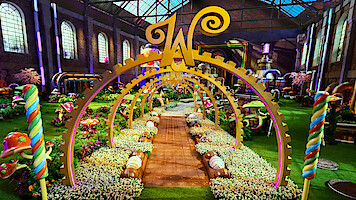 A whimsical candy-themed garden inside a large factory with colorful sweets, oversized mushrooms, lollipops, golden arches, flowers, and a large "W" centerpiece under a glass roof, creating a magical, playful atmosphere.