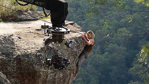 Actor climbing a rocky cliff edge in a forested area, filmed up close by a large movie camera on a crane, during an outdoor film production.