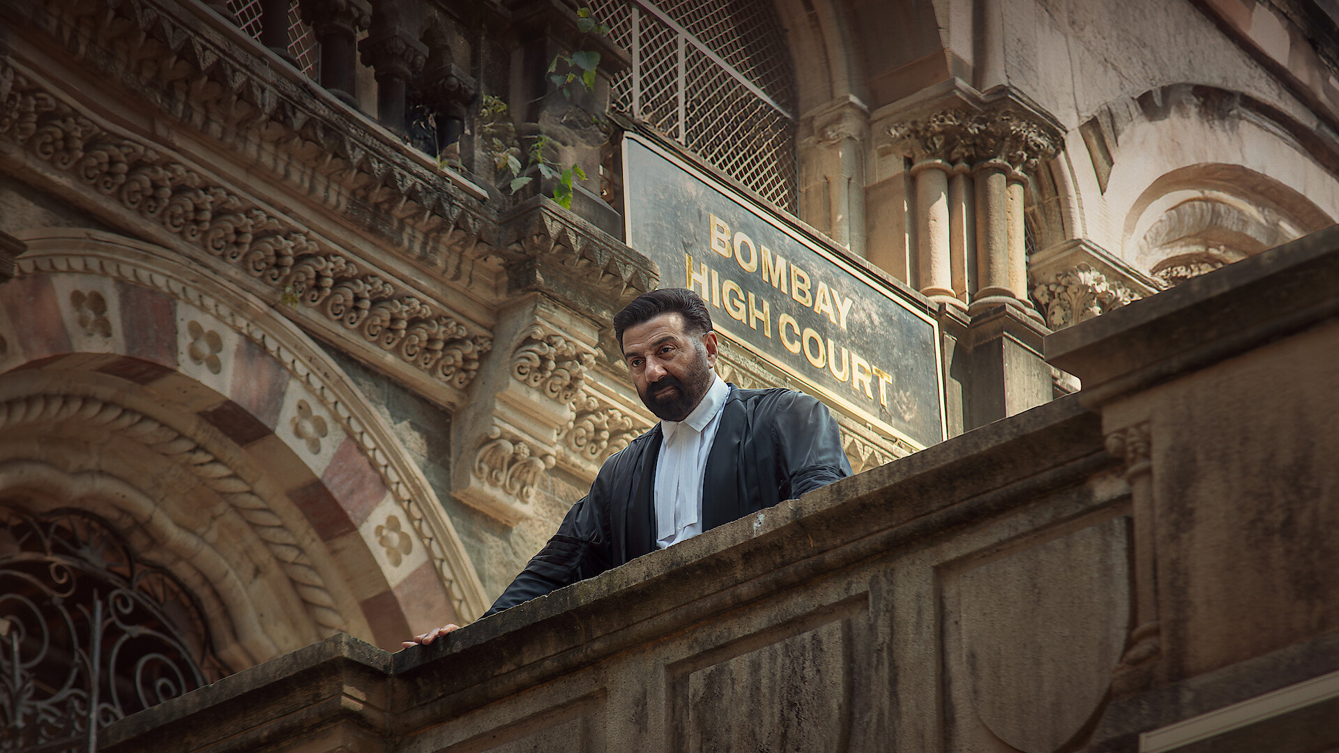 A man in formal attire stands on the steps of an ornate, historic building near a "Bombay High Court" sign, suggesting a legal or courthouse environment.