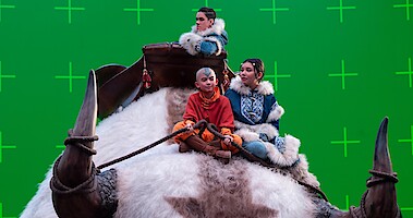A behind the scenes photo of the cast of 'Avatar: The Last Airbender' riding Appa in front of a green screen. 