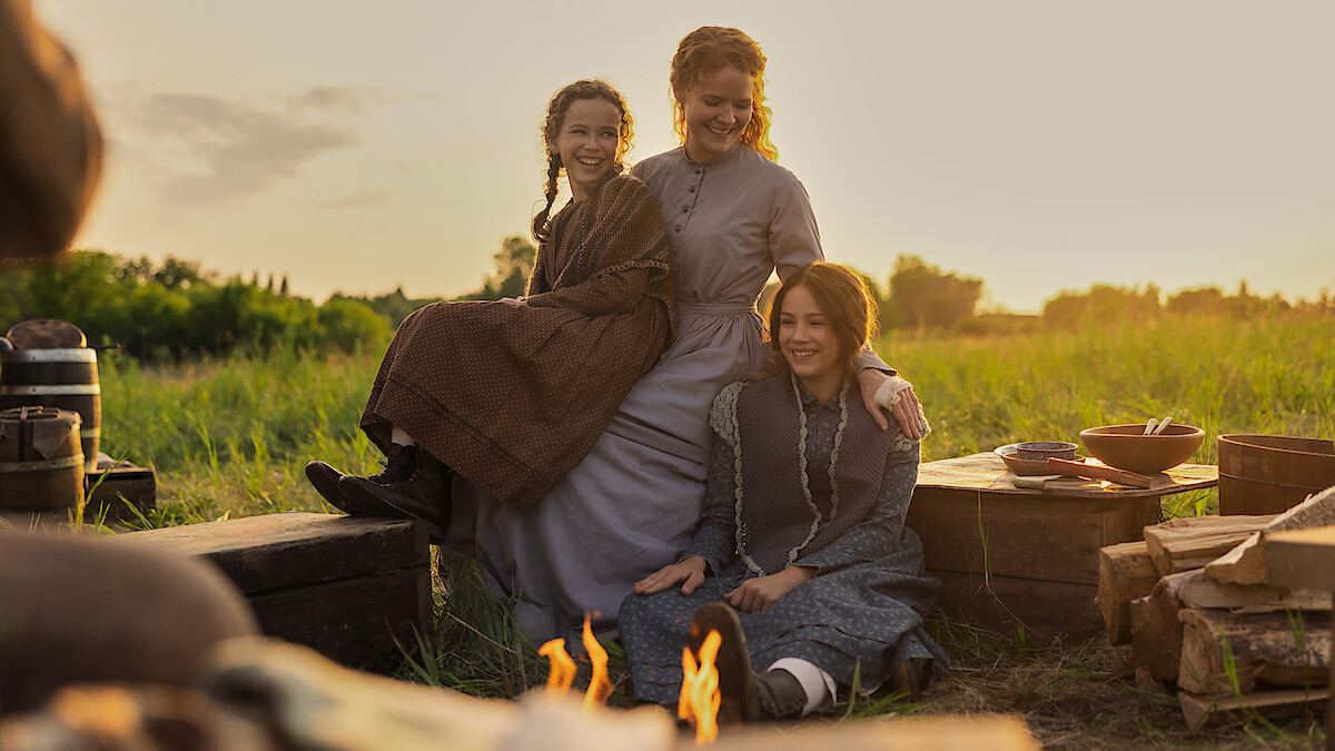 Three women in old-fashioned dresses sitting and smiling outdoors on grass near a campfire, surrounded by wooden crates and cooking utensils in a field at sunset.