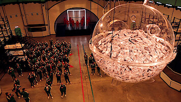 A large group of people in green tracksuits stand in a game arena, looking up at a giant transparent sphere filled with cash, while masked guards in red stand in the background; the mood is tense and competitive.