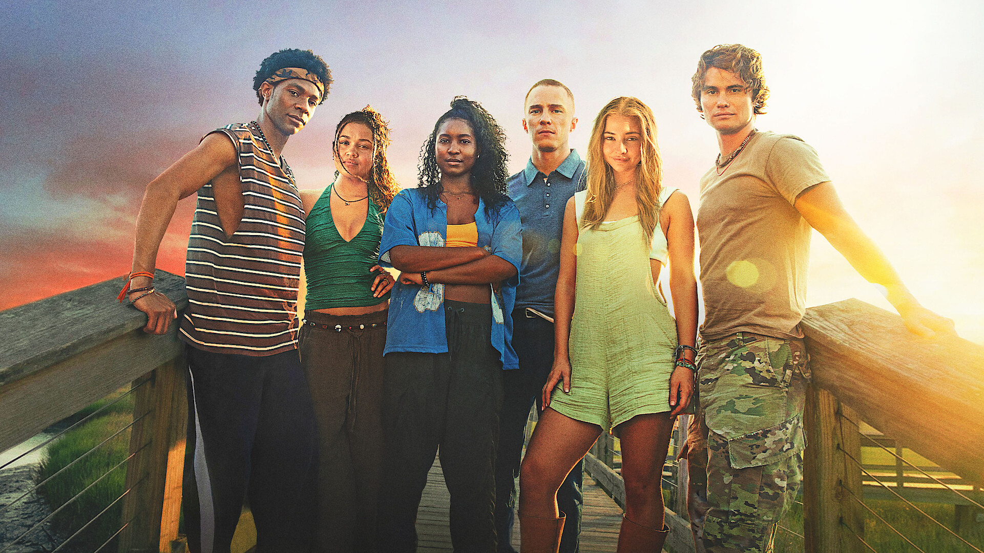 Six young adults stand together on a wooden boardwalk at sunset, with open fields and dramatic skies in the background, posing confidently in casual summer clothing.