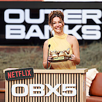 Woman in yellow dress smiling and holding a gold award at a Netflix OBX5 Outer Banks event, standing at a podium with branded signs, outdoor setting with greenery and large logo background.