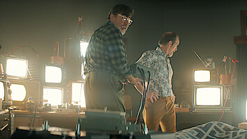 Two men in a dimly lit room filled with old televisions and electronic equipment, one holding a device, standing beside a bed with a patterned blanket, suggesting an experimental or scientific setting.