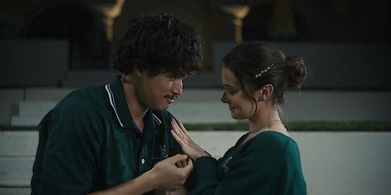 A young man and woman share a tender moment, holding hands and smiling at each other, outdoors at night with white steps and arches in the background, suggesting a romantic or intimate setting.