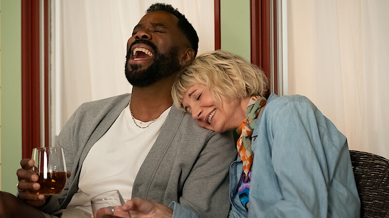 Two people sitting closely together indoors, laughing and smiling. One person is holding a drink while the other leans on their shoulder, creating a relaxed and joyful atmosphere. Warm lighting and comfortable setting.