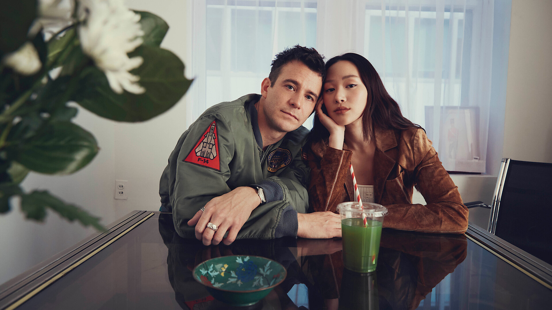 A man and woman sit closely at a glass table in a bright room with large windows. There is a green drink, a blue dish, and a vase of flowers on the table, creating a cozy, intimate indoor setting.