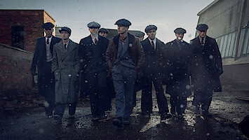 Eight men in vintage suits and flat caps walk together on a wet, gritty street in an industrial urban area, with old brick and metal buildings in the background under a gray, overcast sky.