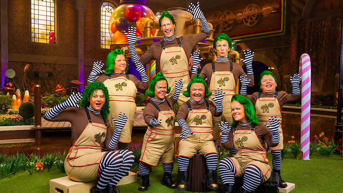 Group of people dressed as Oompa Loompas with green wigs, brown shirts, striped leggings, and overalls in a colorful, whimsical candy factory setting with oversized sweets and bright lighting.