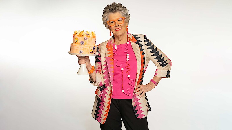 Older woman with gray hair, wearing colorful geometric jacket and glasses, stands in a bright studio holding a decorated cake on a stand, with a confident pose and cheerful expression against a plain white background.