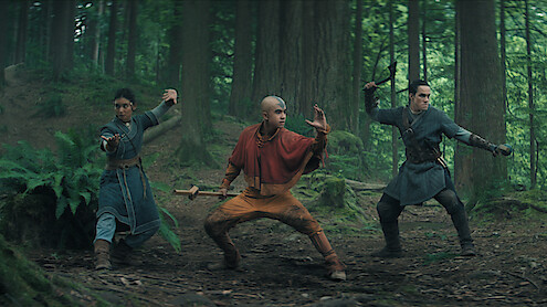 Three people in martial arts stances, dressed in fantasy costumes, stand ready for action in a lush, green forest with tall trees and ferns, suggesting a dramatic, adventurous scene.