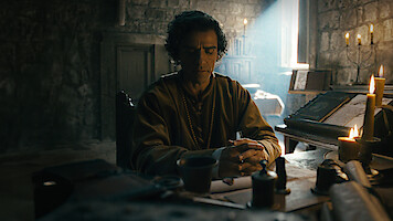 A man sits at a medieval wooden desk in a dimly lit stone room, surrounded by candles, books, and writing tools, with sunlight streaming through a small window, creating a contemplative atmosphere.