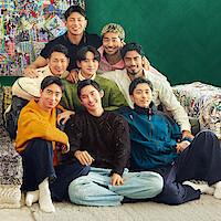 The cast of 'The Boyfriend' Season 2 sit closely together on a colorful sofa in a cozy living room with eclectic pillows, patterned furniture, and a vibrant art piece on a green wall, smiling and posing for the photo.