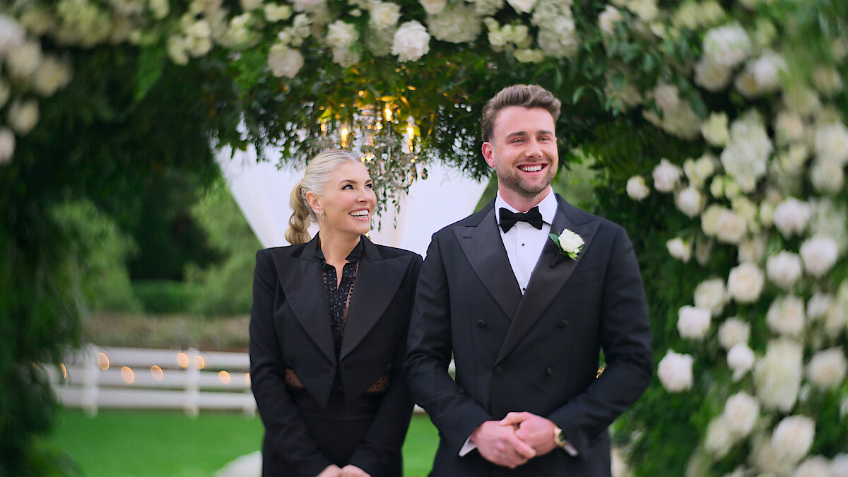 A smiling man in a tuxedo and a woman in formal attire stand under a floral arch outdoors, surrounded by green foliage and white flowers, possibly at a wedding or formal event.