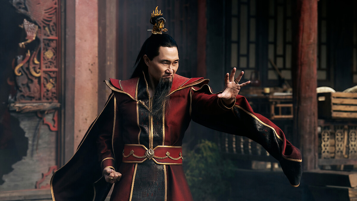 A man in an ornate red robe and crown poses with an outstretched hand in a dramatic stance, standing in a traditional Asian architectural setting with wooden pillars and stone accents in the background.