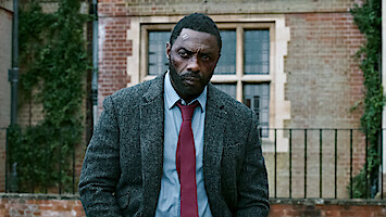 Serious man in a suit with a head injury stands outside a brick building with ivy, giving a tense and somber mood.